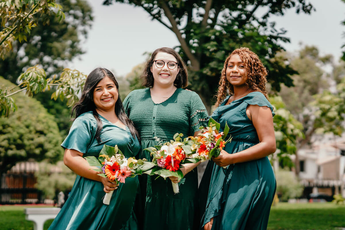 Three bridesmaids with colorful bouquets and green dresses standing in front of some trees.